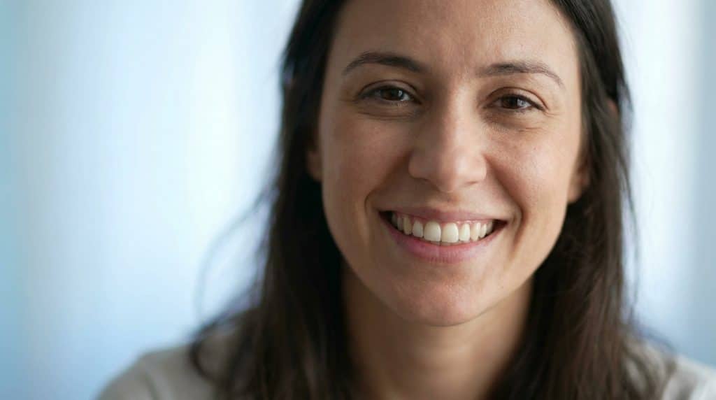 Gros plan d'une femme aux cheveux bruns et aux yeux foncés, souriante, montrant des dents saines sur fond bleu flou.